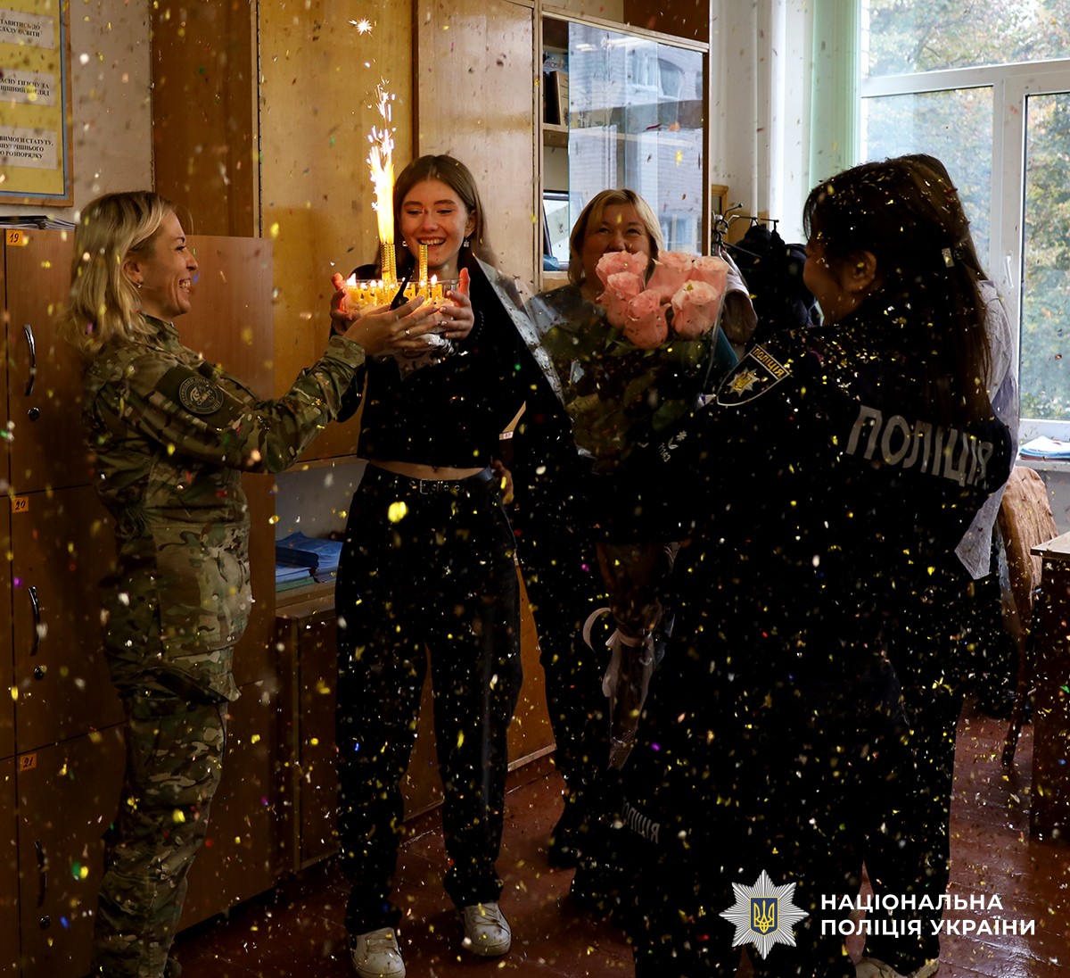 ліворуч у камуфляжі поліцейська тримає торт зі свічкою, яку задуває іменинниця. Поруч із дівчинкою стоять вчителька та ще одна поліцейська з квітами й подарунком.
В повітрі літають блискітки від хлопавки.