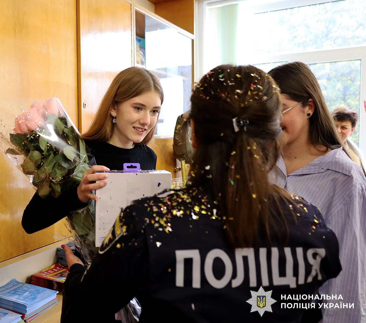 Поліцейська стоїть спиною до камери та вручає подарунок іменинниці. Дівчинка приймає подарунок і тримає в руках квіти. На плечах і волоссі поліцейської видно блискітки.