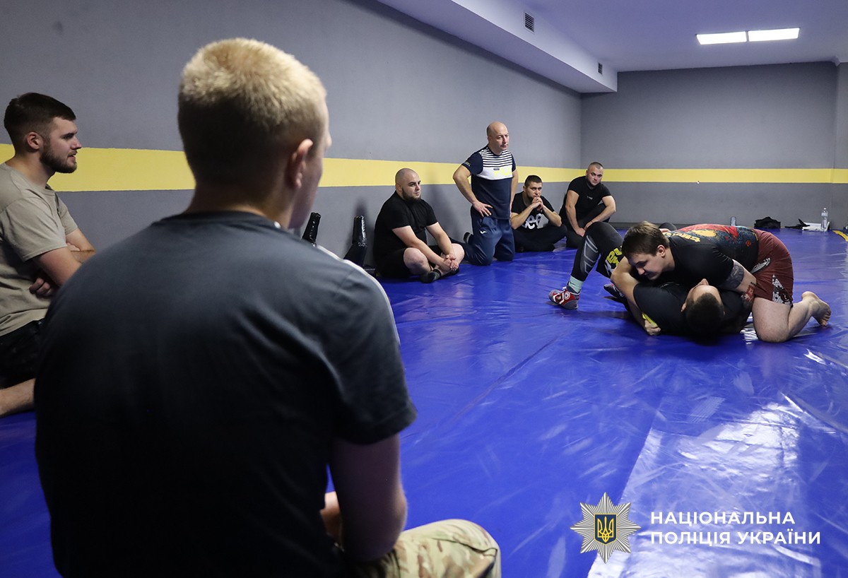 На передньому плані спиною сидить один з учасників, а на задньому плані в колі сидять інші хлопці, у центрі яких тренер демонструє прийом на одному з учасників.
Подія відбувається у спортивному залі.