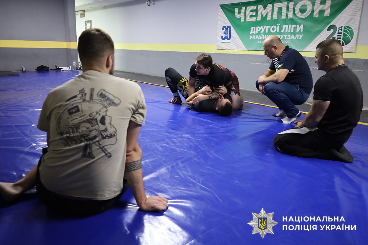 На передньому плані спиною сидить один з учасників, навпроти нього тренер демонструє та роз’яснює прийом на іншому учаснику, а поруч із ним сидить ще один учасник.
Подія відбувається у спортивному залі.