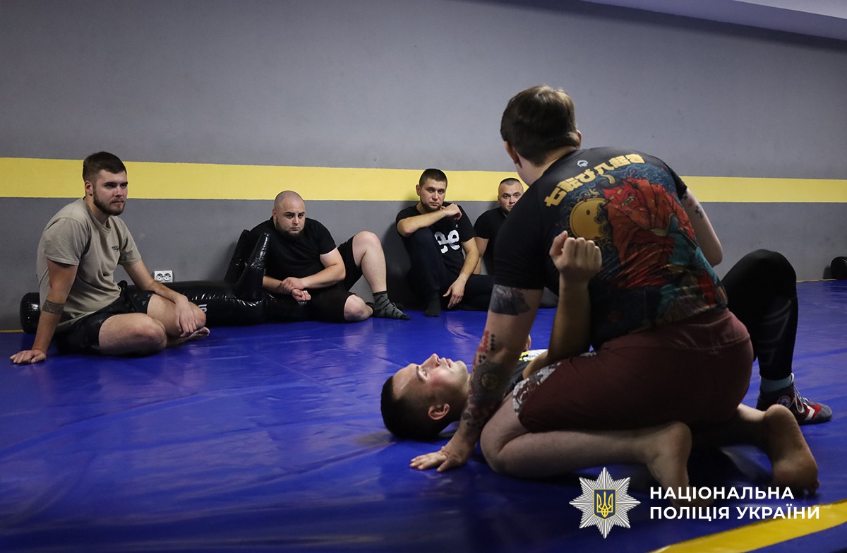 Один з учасників лежить на спині на татамі, тренер показує на ньому прийом, а навпроти сидять інші учасники та уважно спостерігають.
Подія відбувається у спортивному залі.