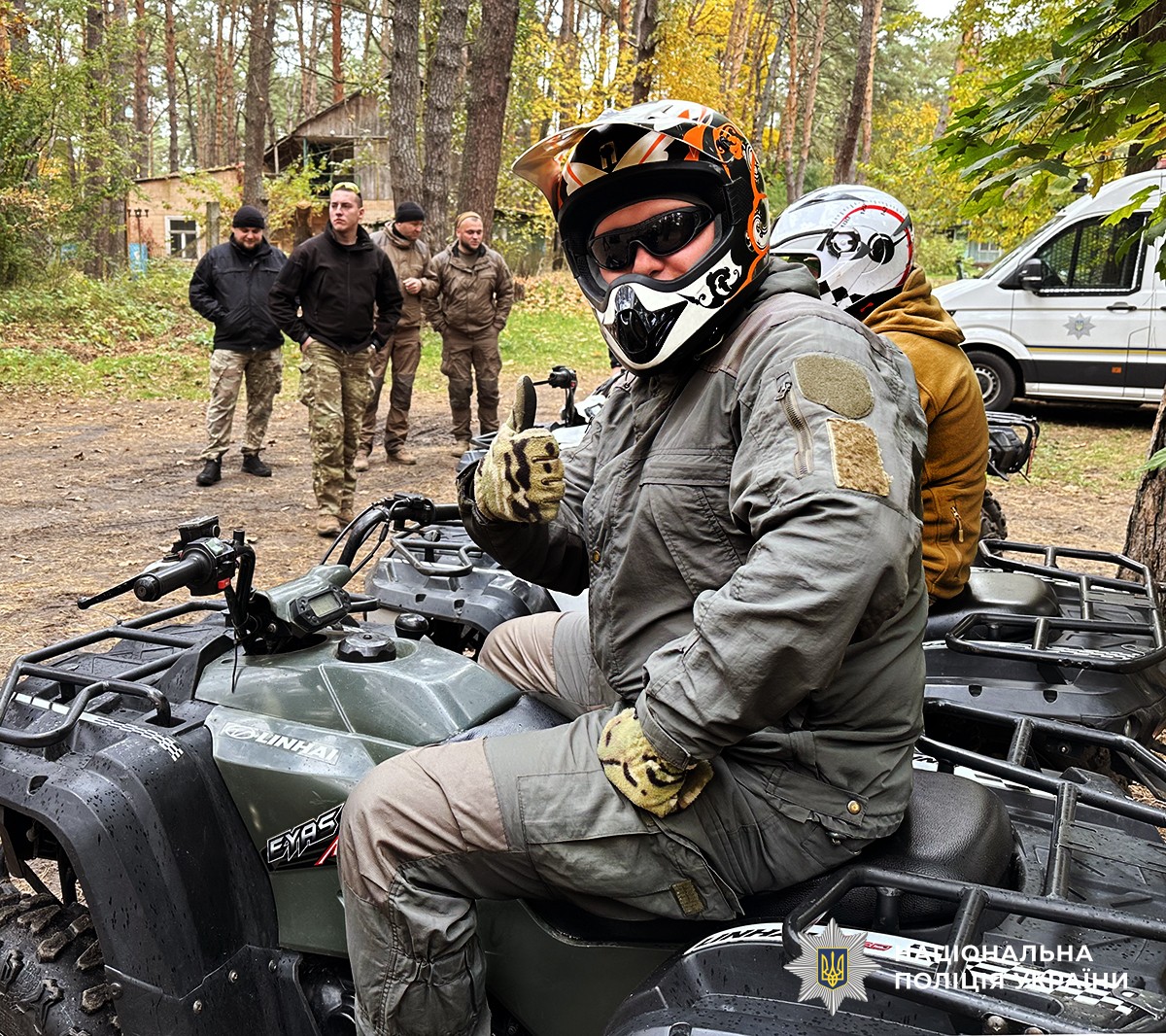 На передньому плані на квадроциклі сидить чоловік у захисному шоломі, який позує для фото. На задньому плані — ще один чоловік на квадроциклі у захисному шоломі, поруч із яким стоять четверо інших чоловіків.
