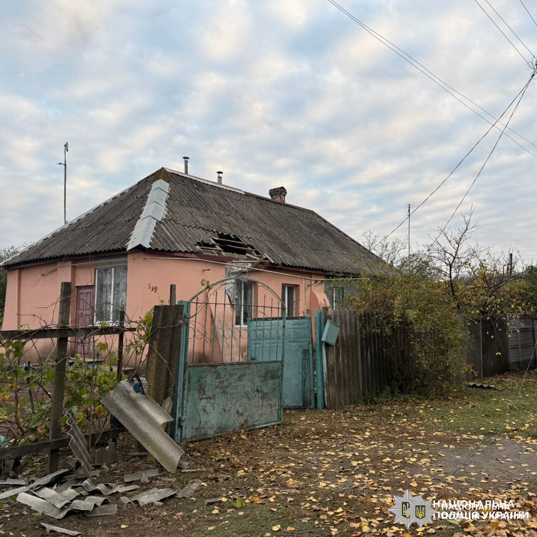 Одноповерховий житловий будинок рожевого кольору з пошкодженим дахом: видно відсутні черепиці або листи покриття. Перед будинком металева брама та паркан, частково відкриті й з ознаками зношеності.