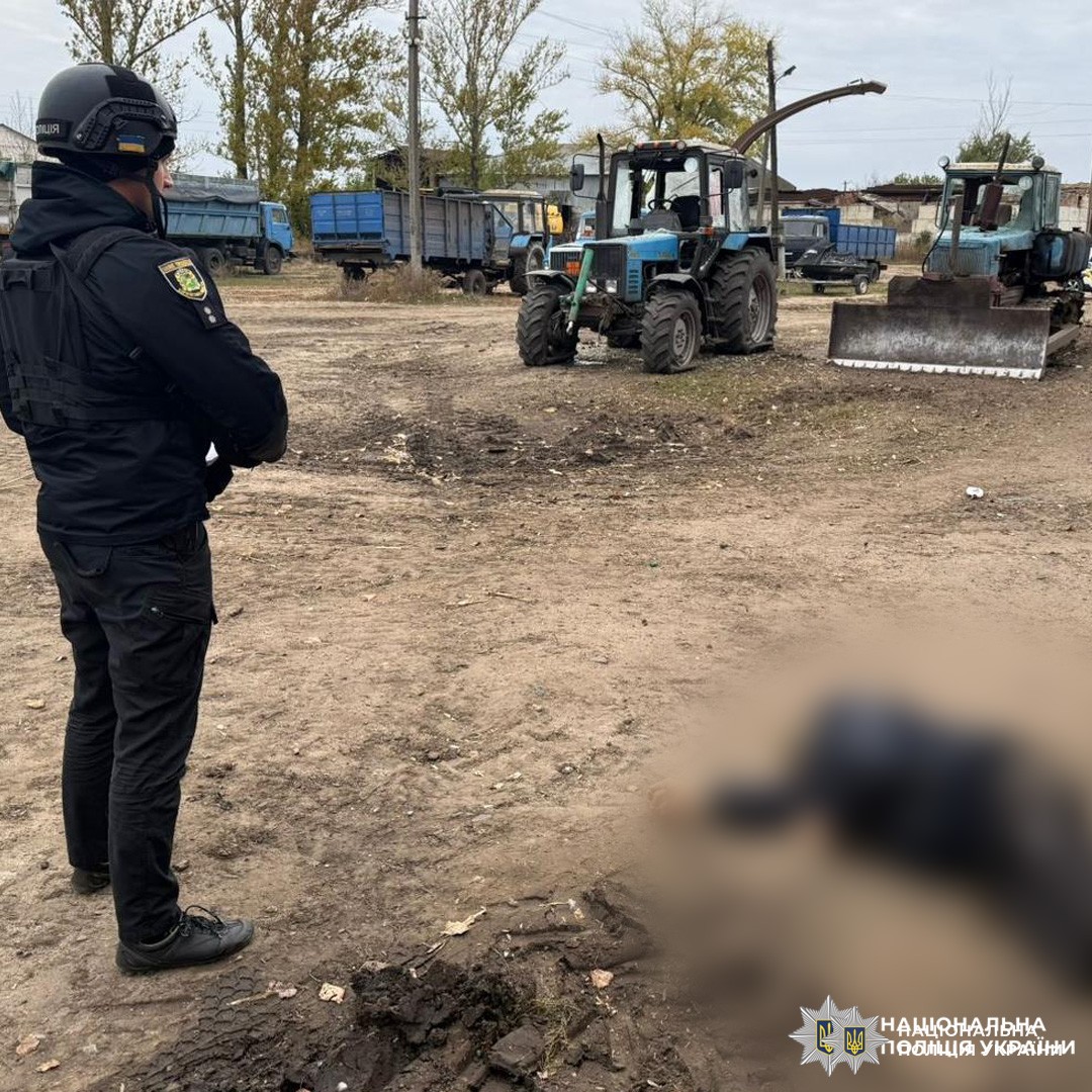 Офіцер у чорній формі та шоломі стоїть на відкритій ділянці з ґрунтовим покриттям.Місце події оточене синіми та зеленими тракторами й вантажівками, що вказує на сільськогосподарське або промислове середовище. На фоні дерева та будівлі.