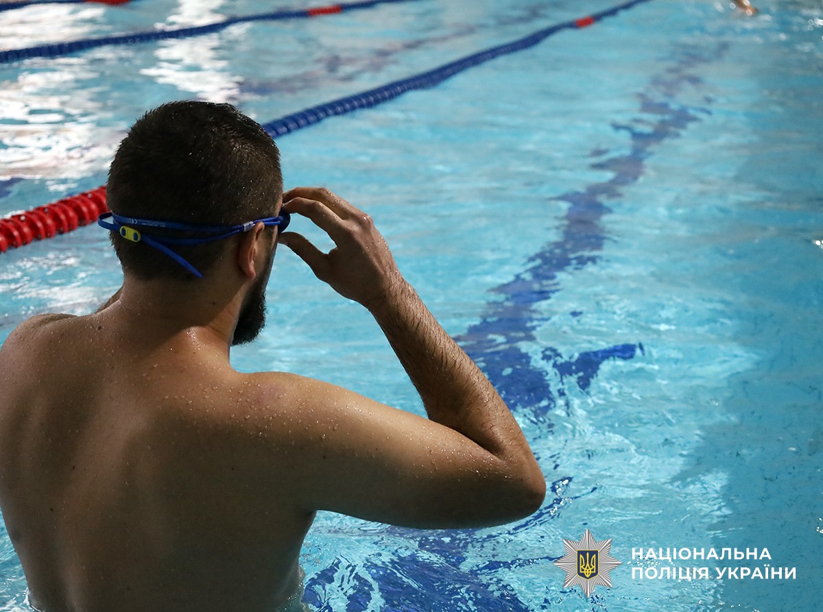 На зображенні чоловік стоїть у басейні спиною до камери. Він готується до запливу, поправляючи свої блакитні окуляри для плавання. Вода довкола чиста й прозора, у басейні видно розділові доріжки синього та червоного кольору.