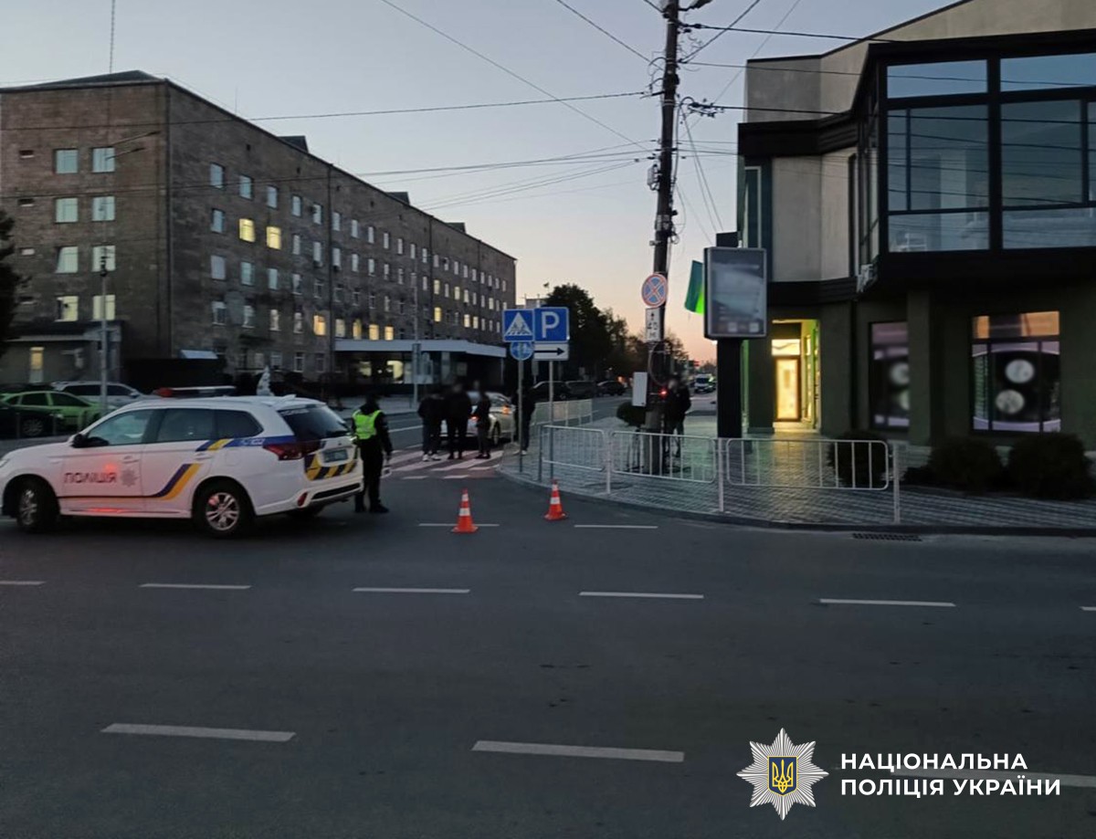 Перехрестя в місті, з обох сторін дороги будівлі, поліцейська машина на дорозі, люди на пішохідному переході