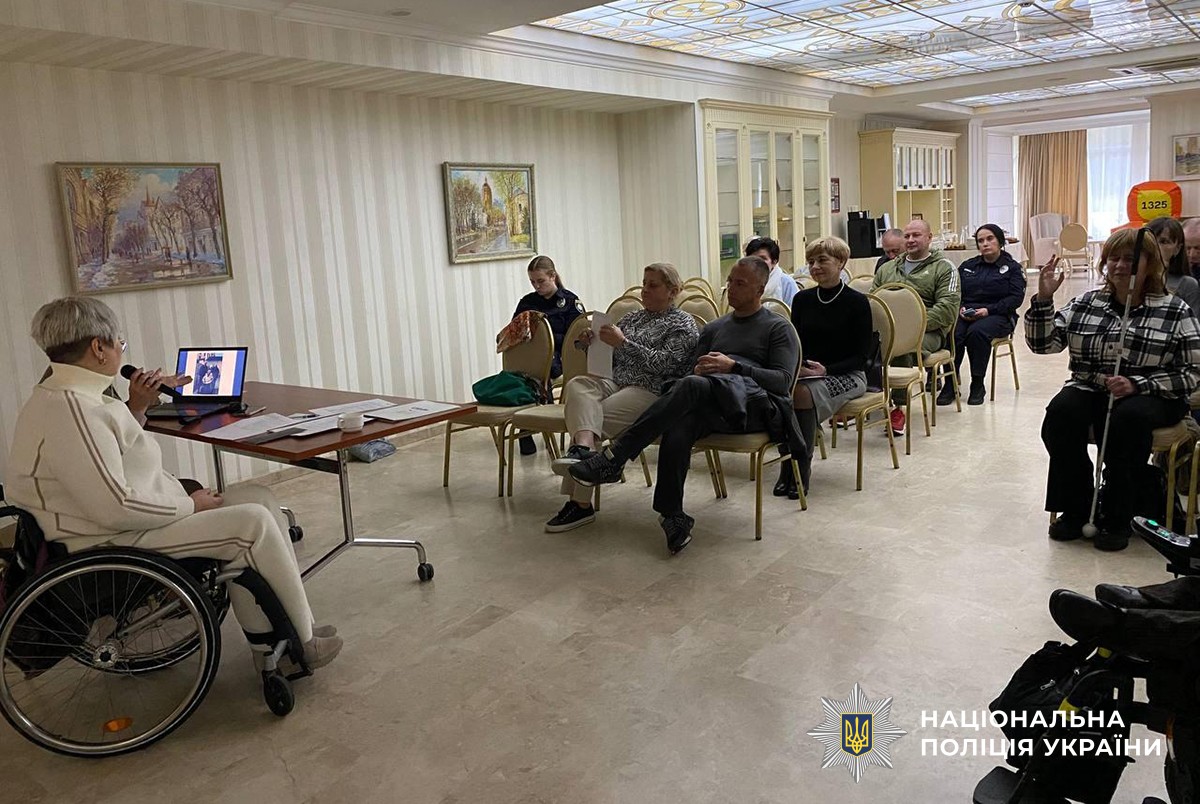 Жінка на кріслі колісному розповідає учасникам зустрічі, які сидять на стільцях в залі