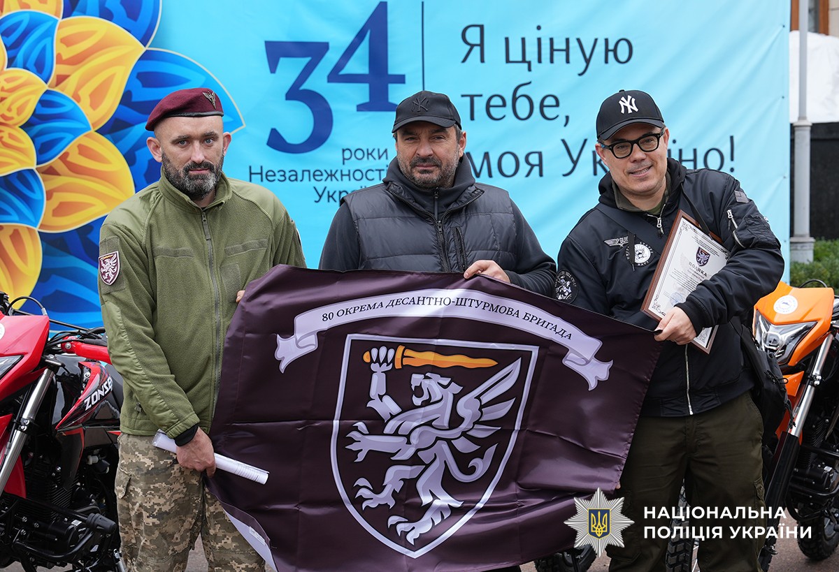 Військовослужбовець, представник благодійної організації та благодійник позують на спільному фото на фоні прапора бригади бордового кольору.