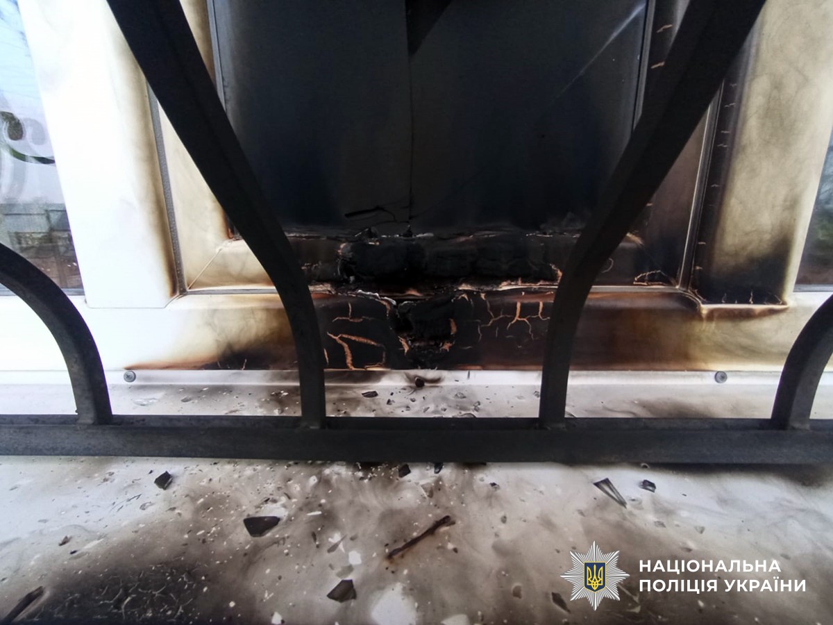 Вікно пошкоджене вогнем: рама почорніла, видно обгорілу ділянку під ним. Перед вікном — металева решітка. На поверхні під рамою — уламки скла та сліди гару.