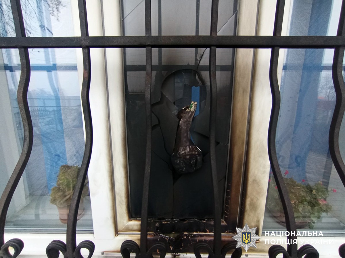 Вікно пошкоджене вогнем: рама почорніла, видно обгорілу ділянку під ним. Перед вікном — металева решітка. На поверхні під рамою — уламки скла та сліди гару.