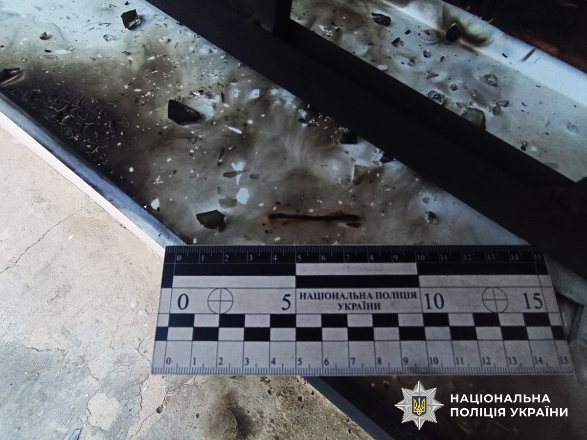 упний план місця події. На поверхні біля вікна видно обвуглені сліди та розсипані уламки скла. На знімку присутня вимірювальна лінійка з маркуванням Нацполіції України.