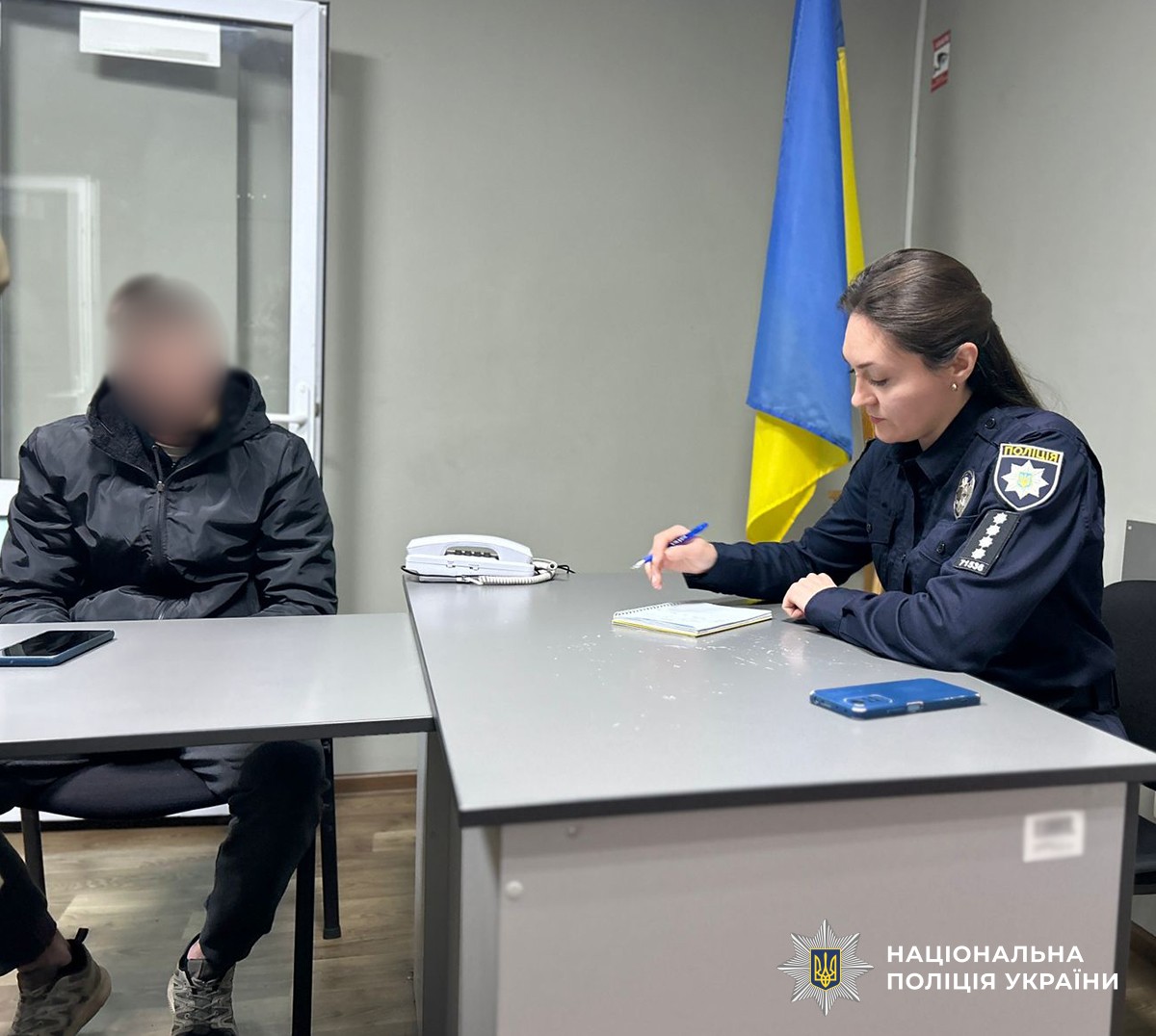 У кабінеті поліції за столом сидять двоє людей. Праворуч — поліцейська, ліворуч — чоловік у темній куртці, обличчя розмите. На столі видно телефон, блокнот та службовий телефон з дротом.