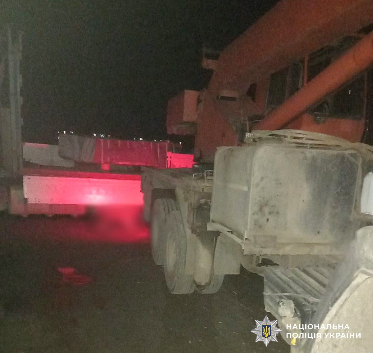 Місце дорожньо-транспортної пригоди, у темну пору доби. Ділянка, де проводилися розвантажувальні роботи: вантажівка-кран та причіп із кам’яними блоками. Місце події освітлене червоним світлом, ймовірно від габаритних вогнів або проблискових маячків. Між транспортними засобами заблюрене тіло людини.