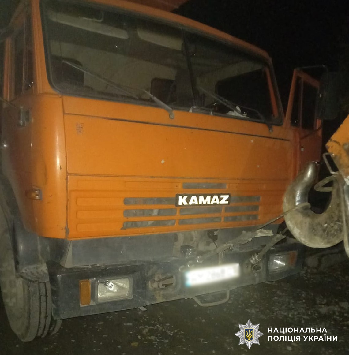 Видно передню частину помаранчевого автокрану КАМАЗ, з відкритими дверцятами кабіни та крановим гаком попереду.
