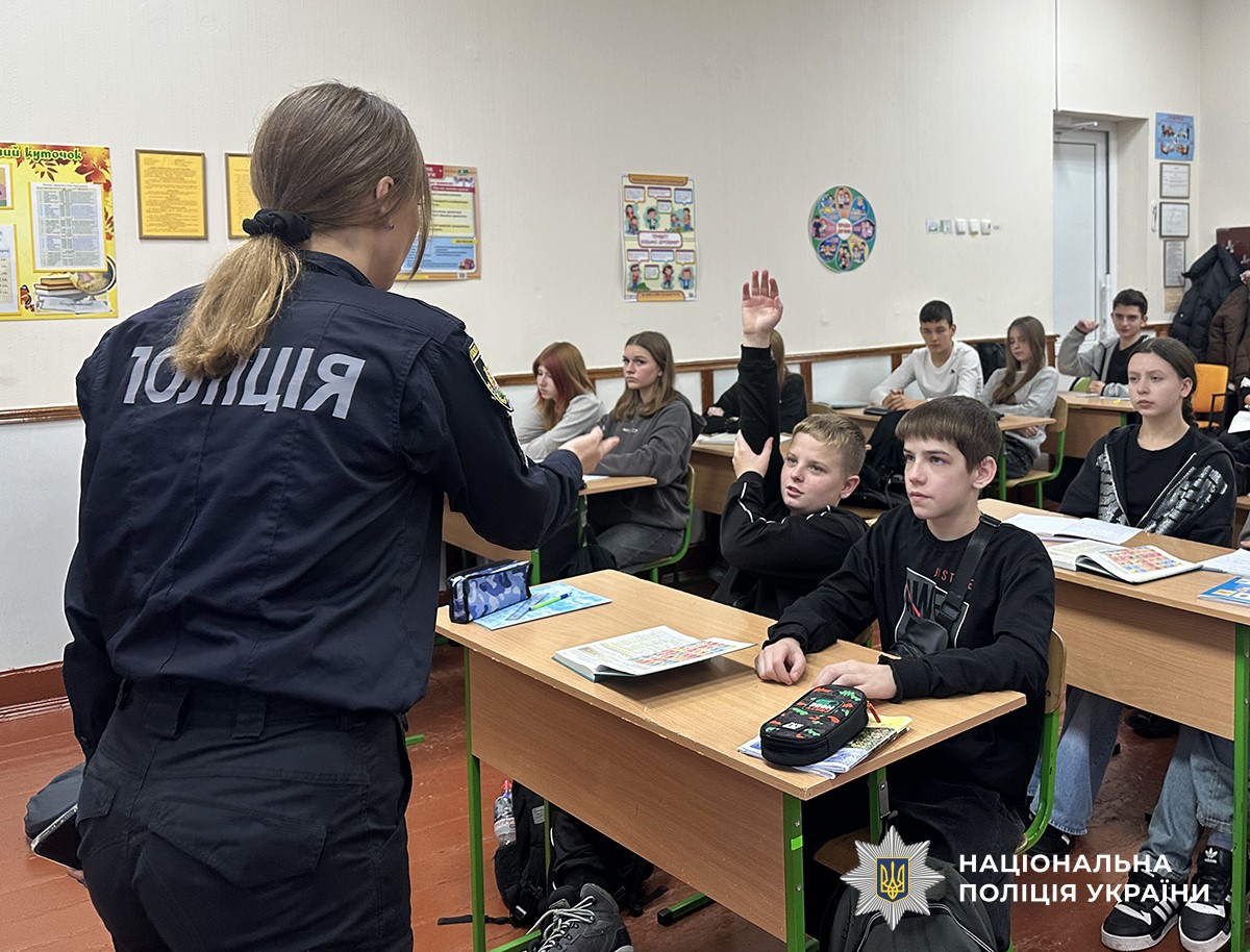 Поліцейська спілкується з учнями у класі. Один хлопець піднімає руку, решта уважно слухають. Атмосфера жвава, діалогова.