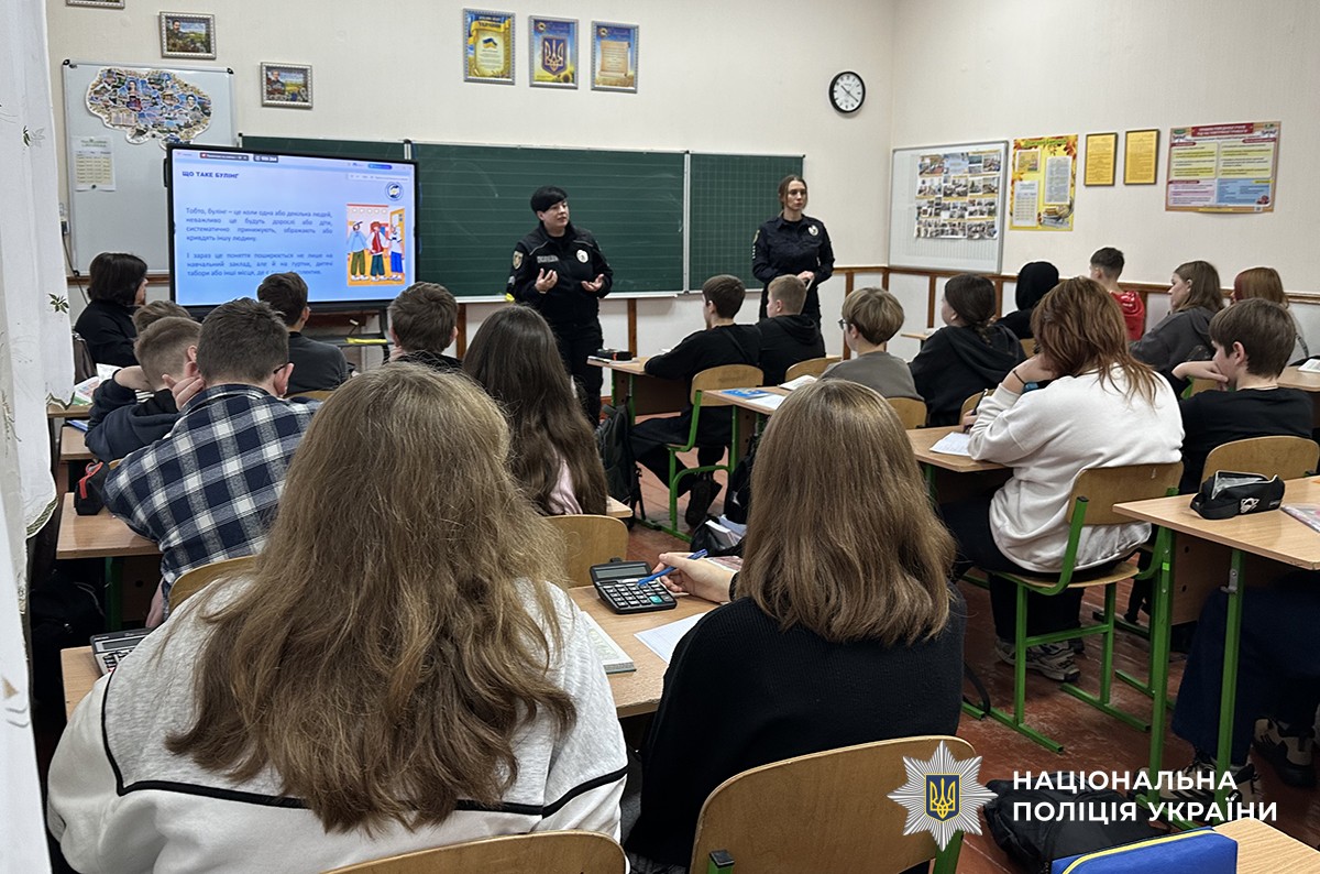 Двоє поліцейських проводять заняття перед класом. На екрані демонструється слайд із текстом про булінг, учні сидять за партами й слухають.