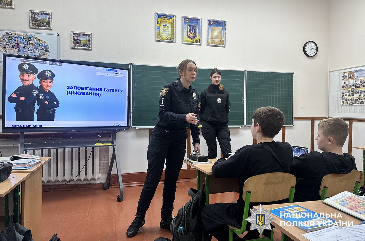 Поліцейська пояснює матеріал біля інтерактивного екрана з темою «Запобігання булінгу (цькуванню)», поруч стоїть її колега. Позаду на стіні селена шкільна дошка.