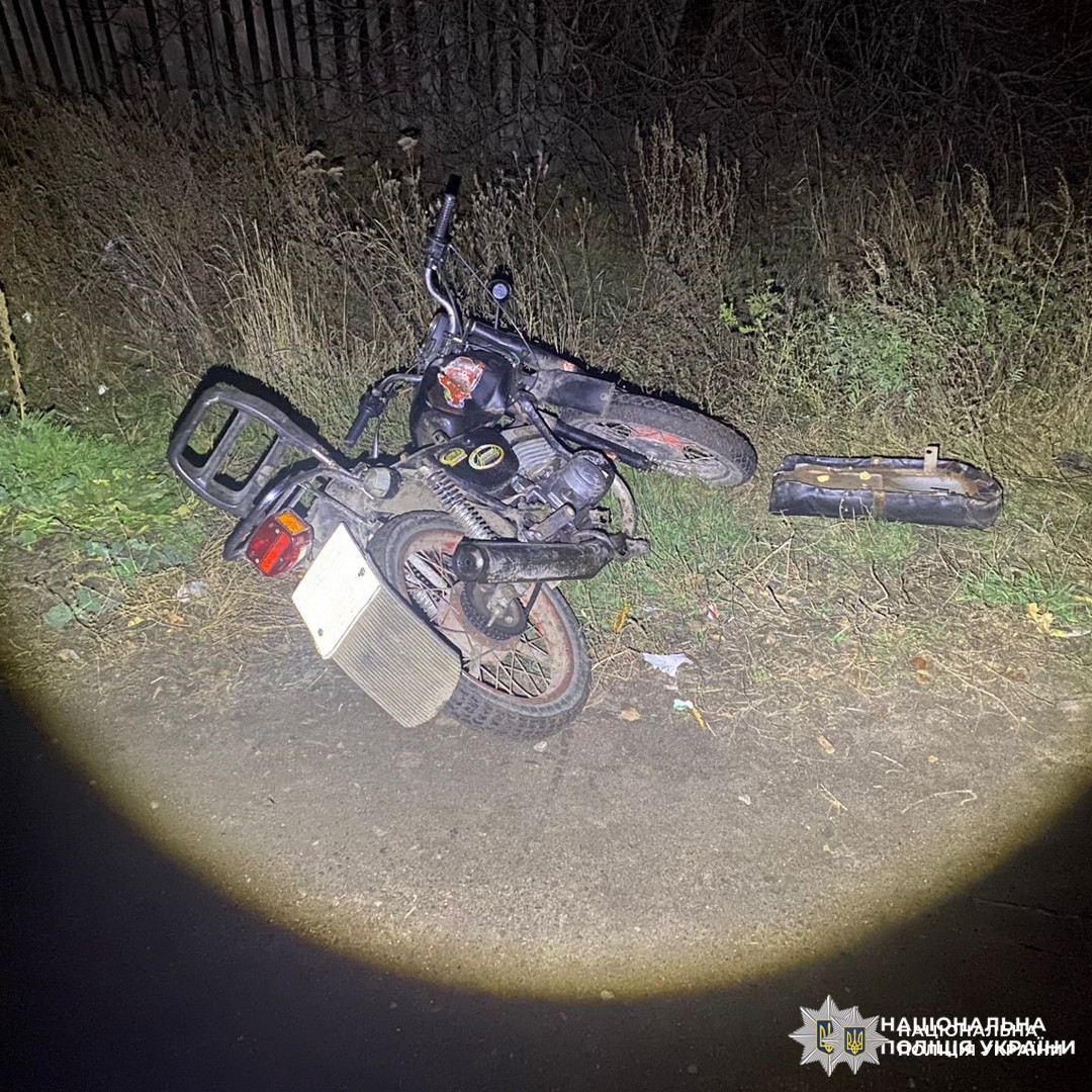 Пошкоджений мотоцикл лежить на боці на узбіччі дороги; поруч відокремлена деталь. Місце події освітлене, у фоні паркан і рослинність.