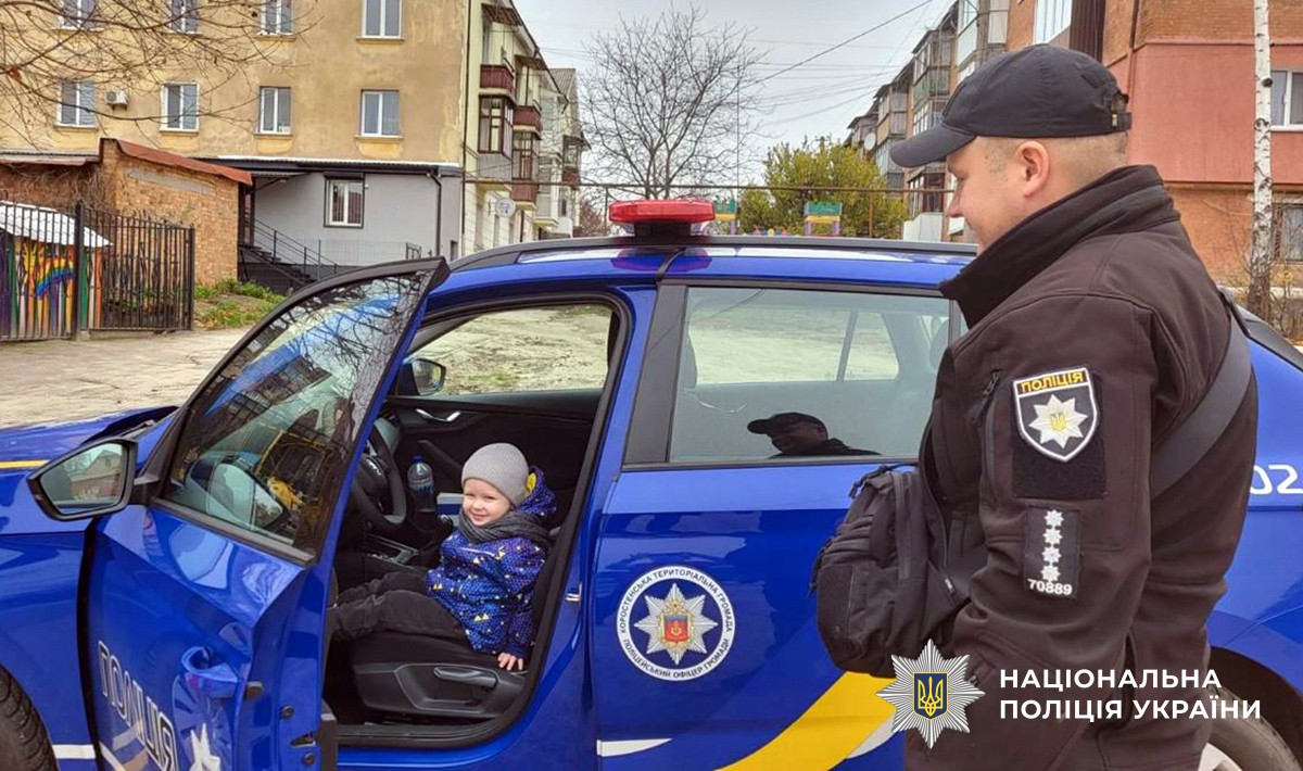 Дитина сидить у поліцейському авто, поруч стоїть поліцейський