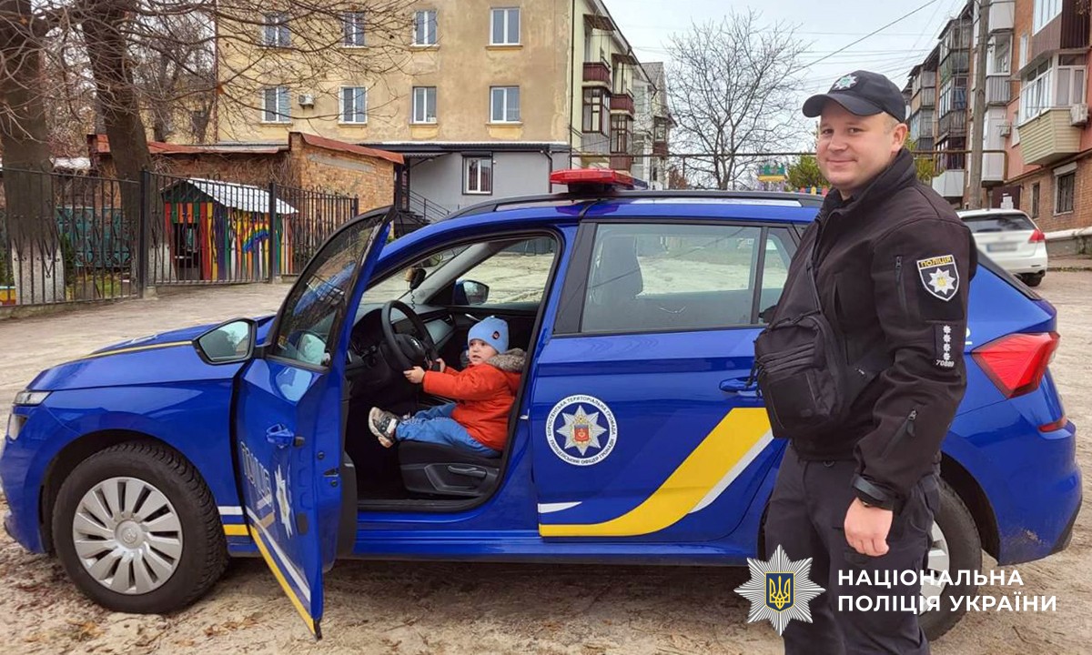 Дитина сидить у поліцейському авто, поруч стоїть поліцейський
