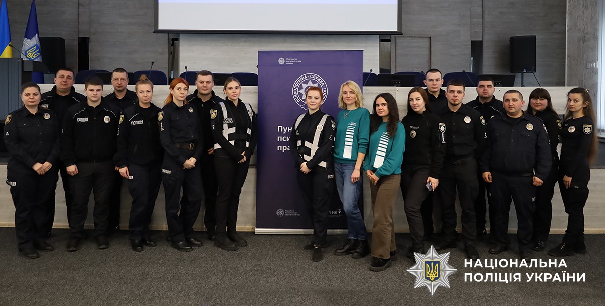 Учасники позують на спільному фото біля трибуни та банера, на якому розміщена емблема та напис «Пункт психологічних практик».