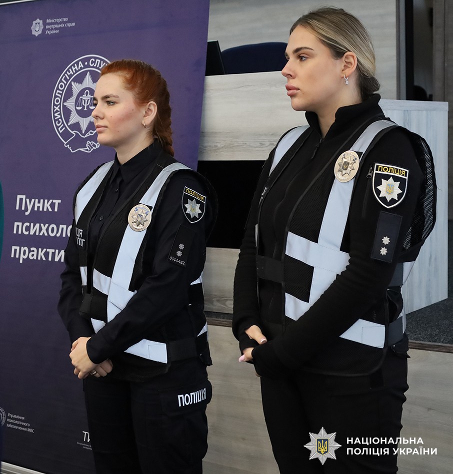 Дві психологині поліції стоять поруч із банером, на якому розміщена емблема та напис «Пункт психологічних практик».