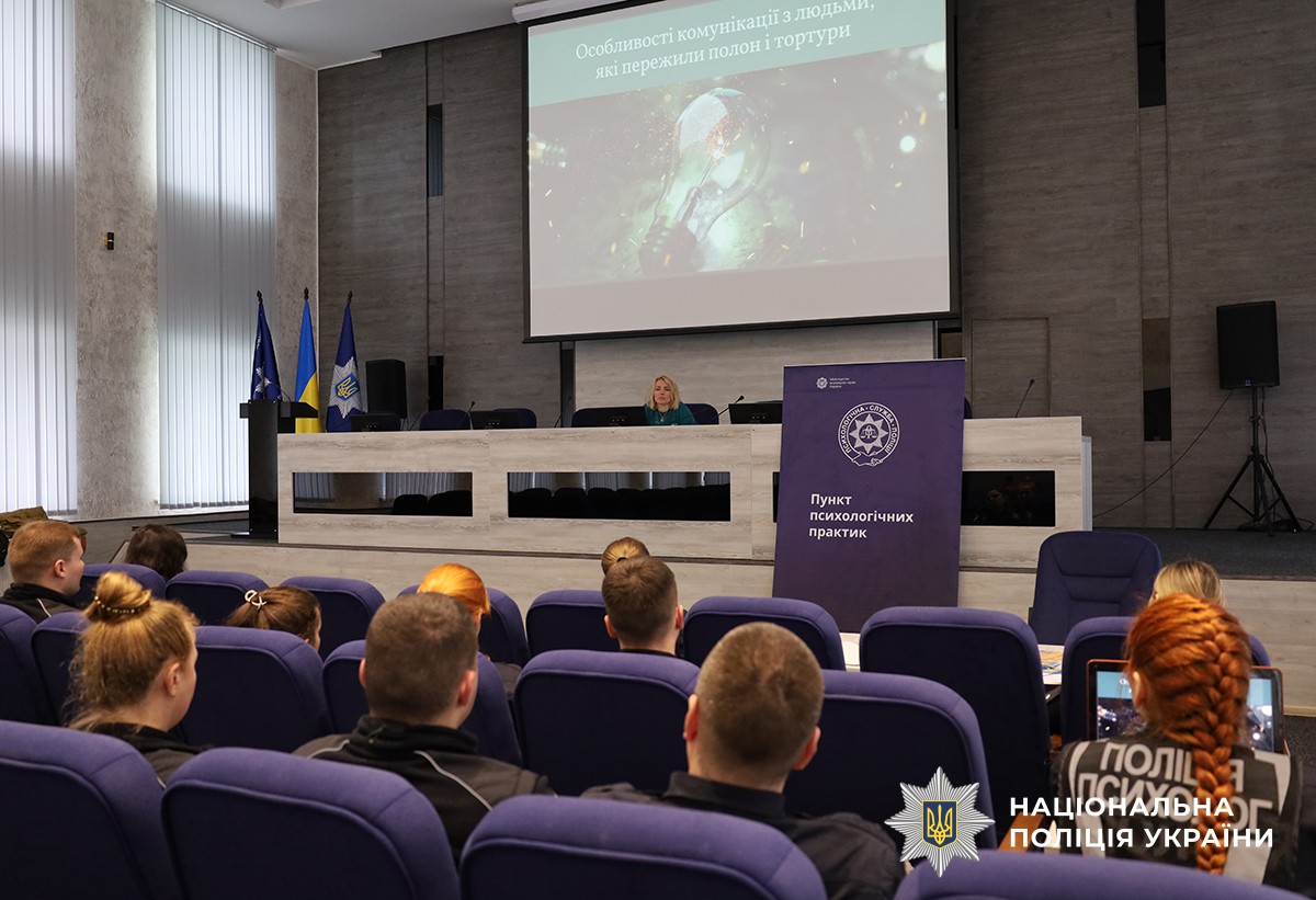 В актовому залі поліцейські слухають психолога, яка виступає за трибуною. На стіні працює проектор, на якому демонструється презентація на тему кризової комунікації. Поряд з трибуною стоїть банер з емблемою та написом «Пункт психологічних практик».