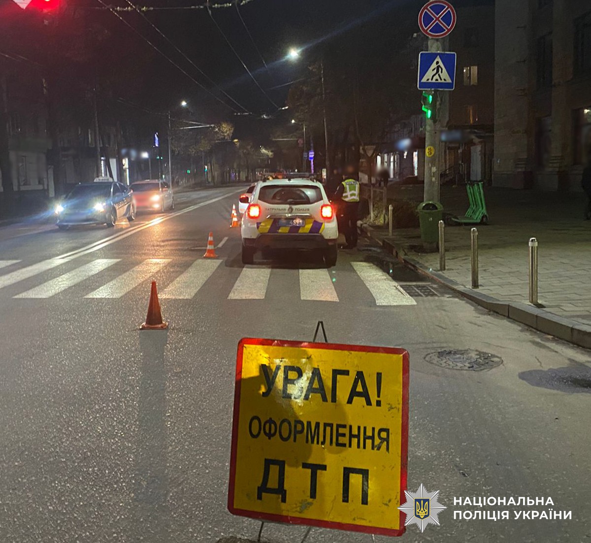 Дорога, освітлена вуличними ліхтарями. На дорозі встановлено жовтий попереджувальний знак «Увага! Оформлення ДТП». Позаду знаку поліцейський автомобіль. Поряд із авто стоїть поліцейський.