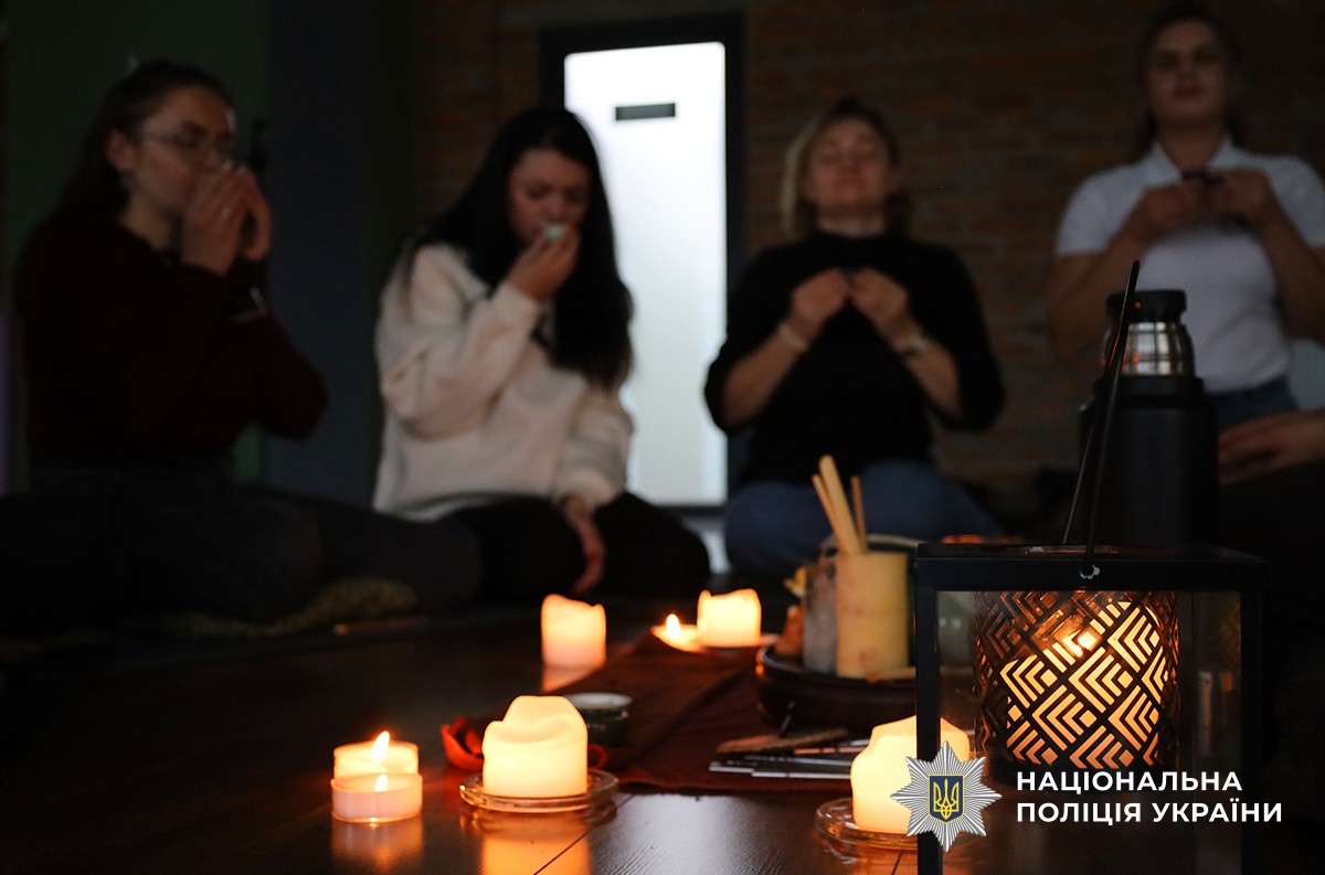 Люди сидять у колі на підлозі зі свічками, тримають чашки, створюючи спокійну, медитативну атмосферу.