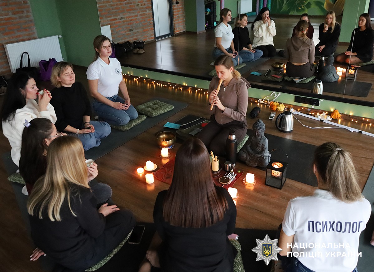 Група людей сидить у колі навколо свічок і статуетки Будди, одна жінка грає на флейті, атмосфера гармонії та релаксу.