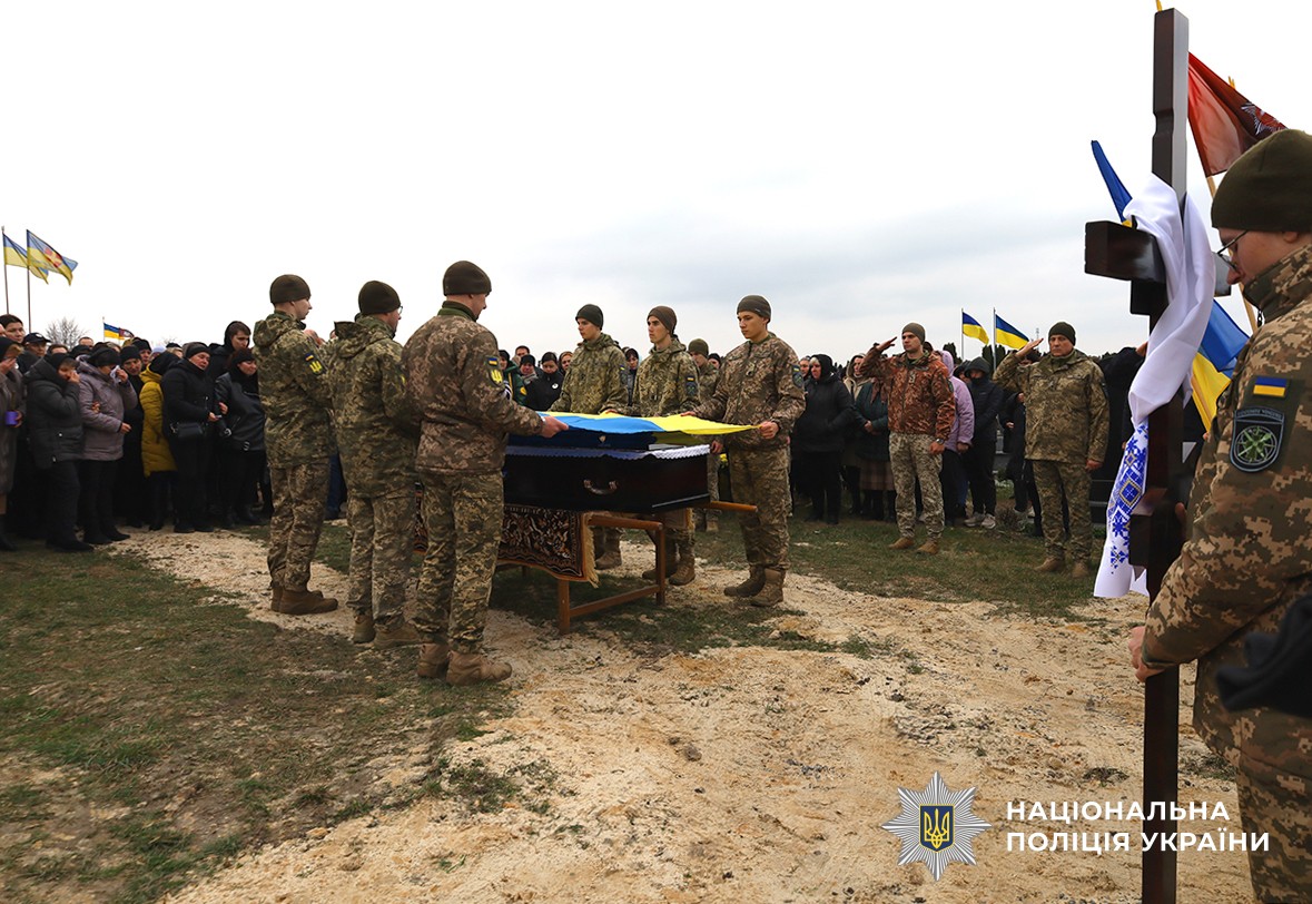 На кладовищі проходить церемонія поховання. Над труною тримають прапор України, навколо зібралися люди та побратими, які віддають честь.