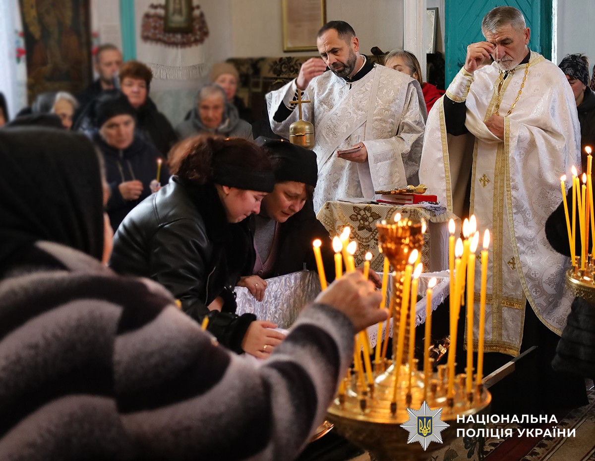 У церкві люди з запаленими свічками стоять на поминальній службі. Мати та дружина загиблого схилились  труною.
