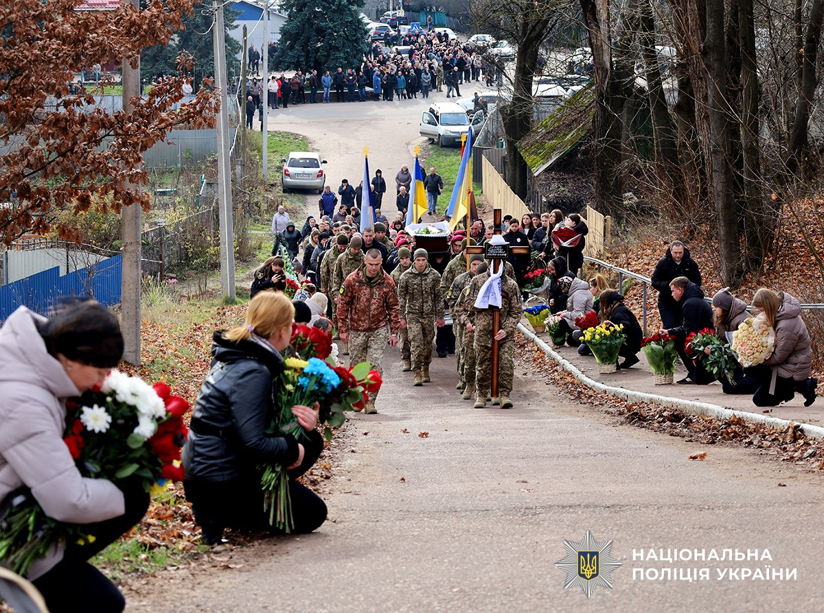 Військові несуть хрест та прапори, за ними поліцейські із труною.  Люди по обидва боки дороги стоять на колінах із квітами, віддаючи шану загиблому.