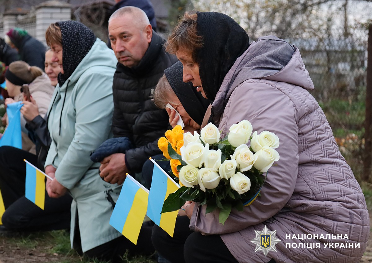 Люди стоять на колінах уздовж дороги, тримаючи  українські прапорці та квіти. На їхніх обличчях скорбота.
