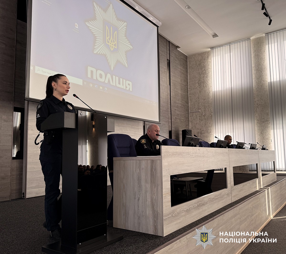 На трибуні на сцені стоїть поліцейська у форменому одязі, поруч за столом сидять двоє поліцейських. На задньому плані екран із зображенням поліцейської зірки та написом «Поліція» на синьому фоні.