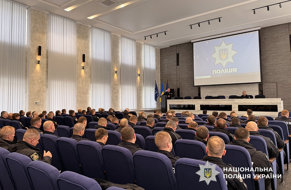 Зал, ракурс іззаду: на синіх стільцях сидять поліцейські, перед ними за трибуною поліцейський щось говорить. На задньому плані видно екран із зображенням поліцейської зірки та написом «Поліція» на синьому фоні.