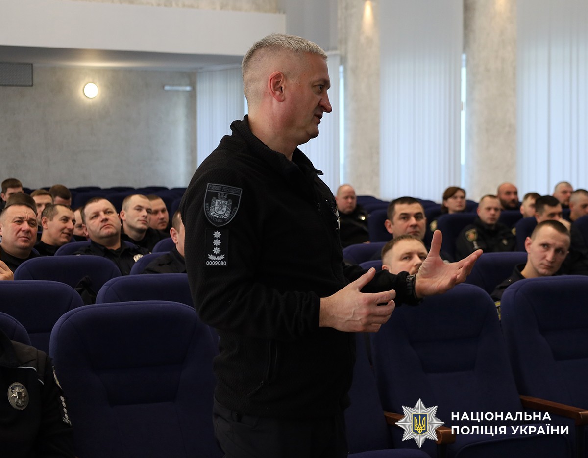 Поліцейський стоїть в актовому залі, говорить і жестикулює руками. На задньому плані на синіх стільцях сидять поліцейські.