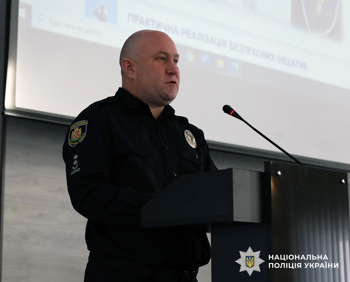 За трибуною стоїть поліцейський. На задньому плані видно екран із демонстрацією інформації.