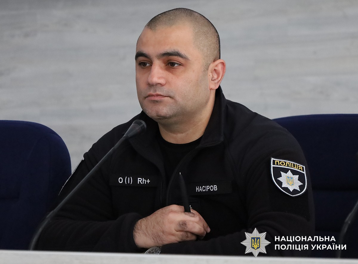 Поліцейський сидить за столом в актовому залі, тримає в руказ ручку.