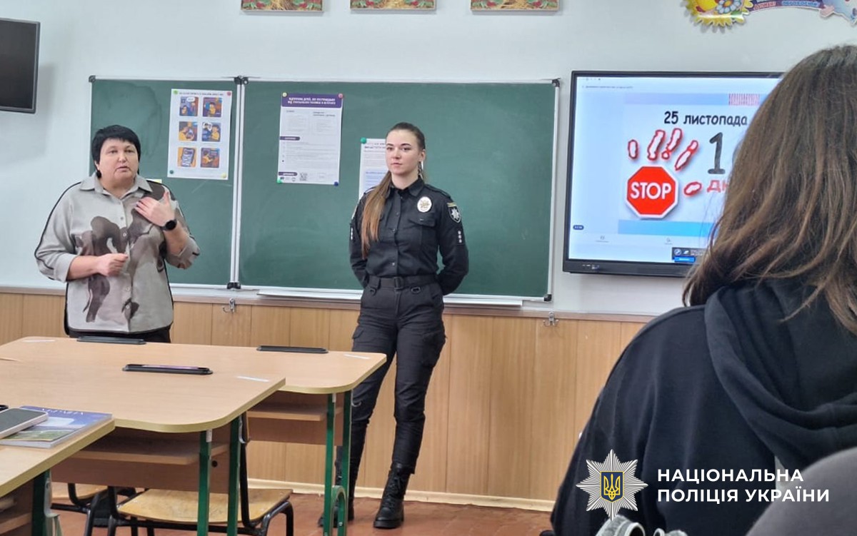 Ювенальна поліцейська стоїть в центрі класу біля шкільної дошки, поруч працівниця соцслужби веде розмову з дітьми