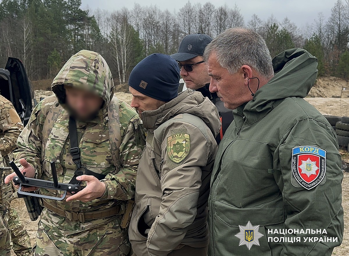 Четверо чоловіків у військовій та тактичній формі на полігоні. Один з них тримає обладнання для керування дроном, інші уважно дивляться.