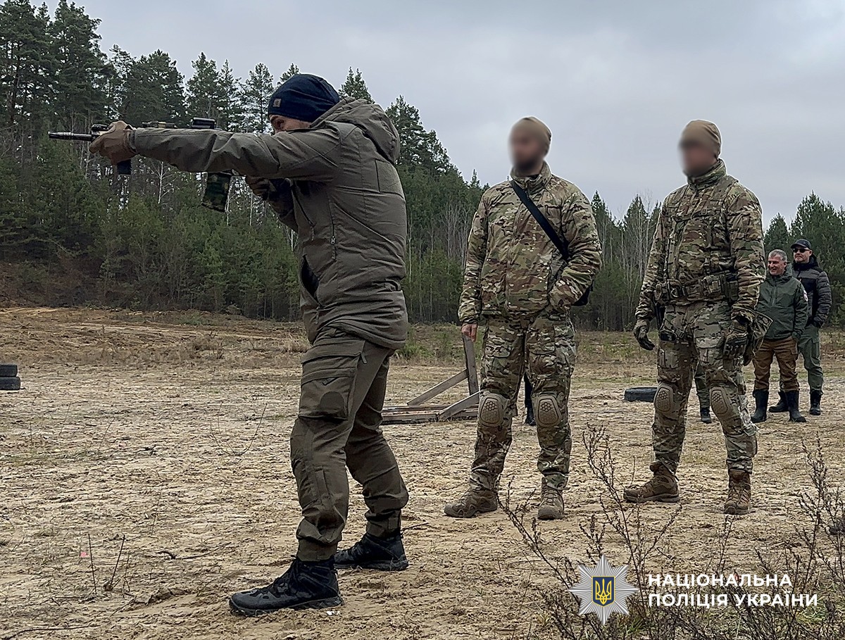 На полігоні троє чоловіків у військовій та тактичній формі. Один із них прицілюється зі зброї, а решта спостерігають за процесом.
