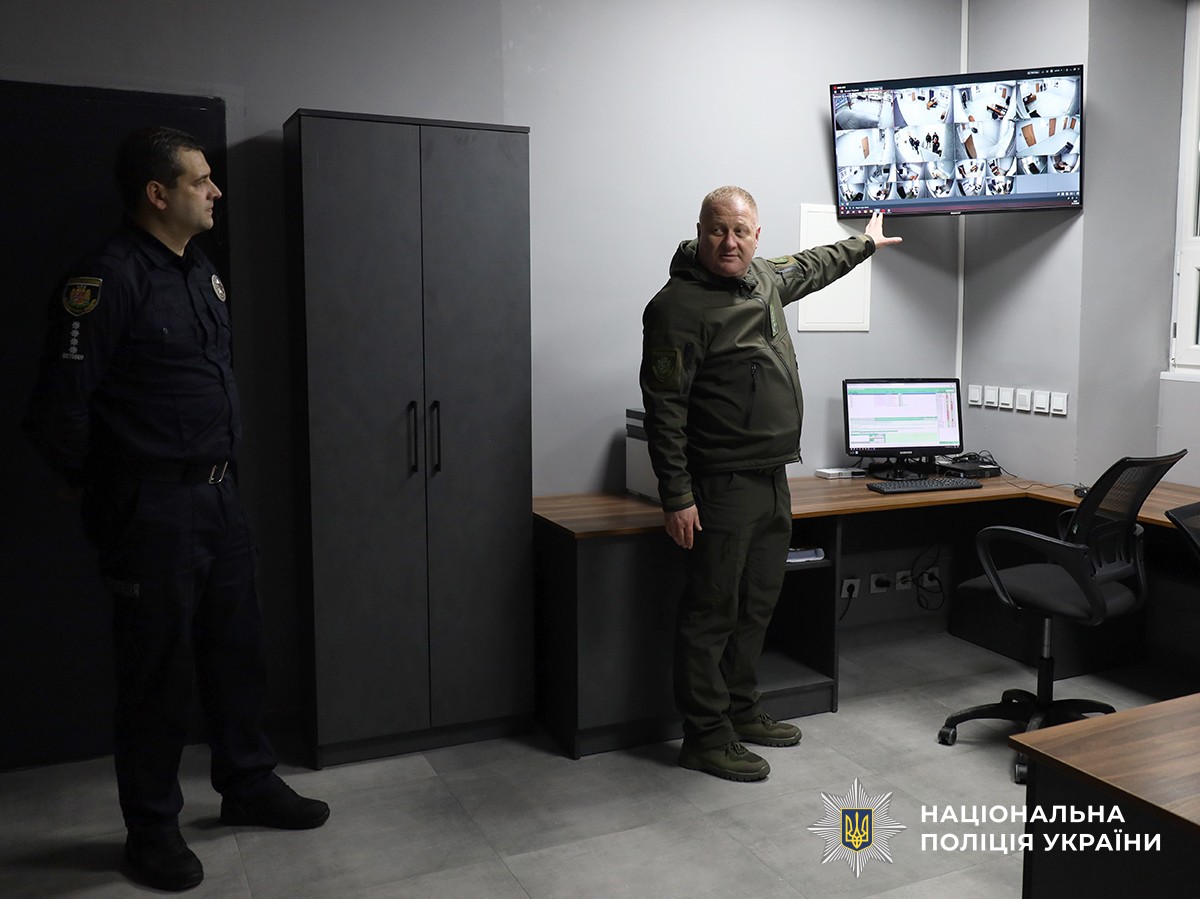 У приміщенні поліції поліцейський жестом указує на екран, на якому виведене зображення з камер відеоспостереження. Поруч із ним стоїть ще один поліцейський.