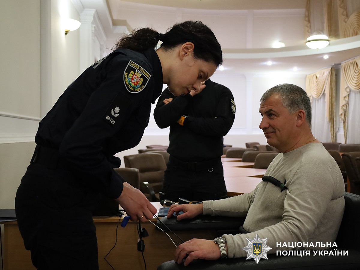 У конференц-залі поліцейська надягає на представника з Естонії обладнання для детектора брехні для презентаці. Позаду них стоїть ще один поліцейський.