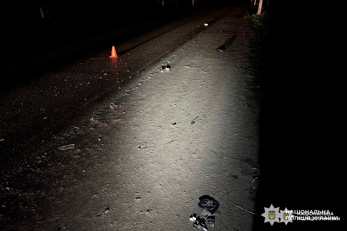 Нічна ділянка дороги з розкиданими дрібними уламками на асфальті та узбіччі. Місце позначене помаранчевим сигнальним конусом, освітлене штучним джерелом світла.