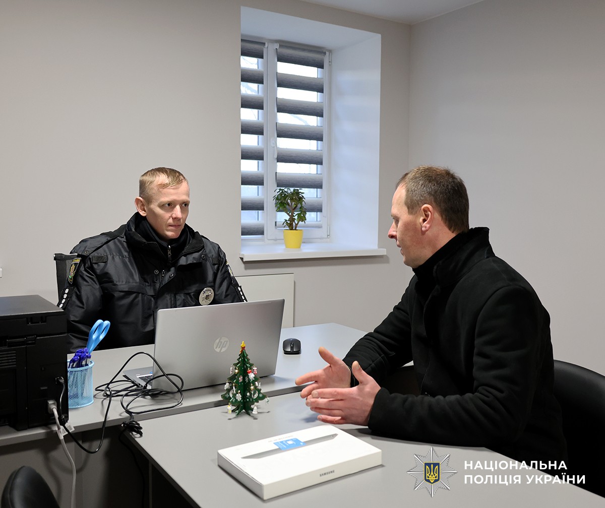 Поліцейський в кабінеті, сидить за столом та слухає громадянина, який сидить за столом навпроти нього.