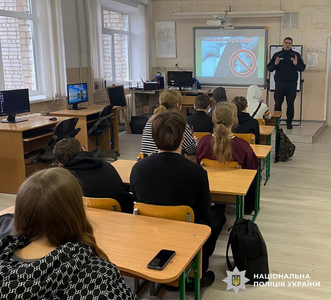 У шкількому класі, діти сидять за партами, навпроти них стоїть поліцейський та щось розповідає жистикулюючи руками. Поруч з поліцейським екран з демонтрацією відеоролика.