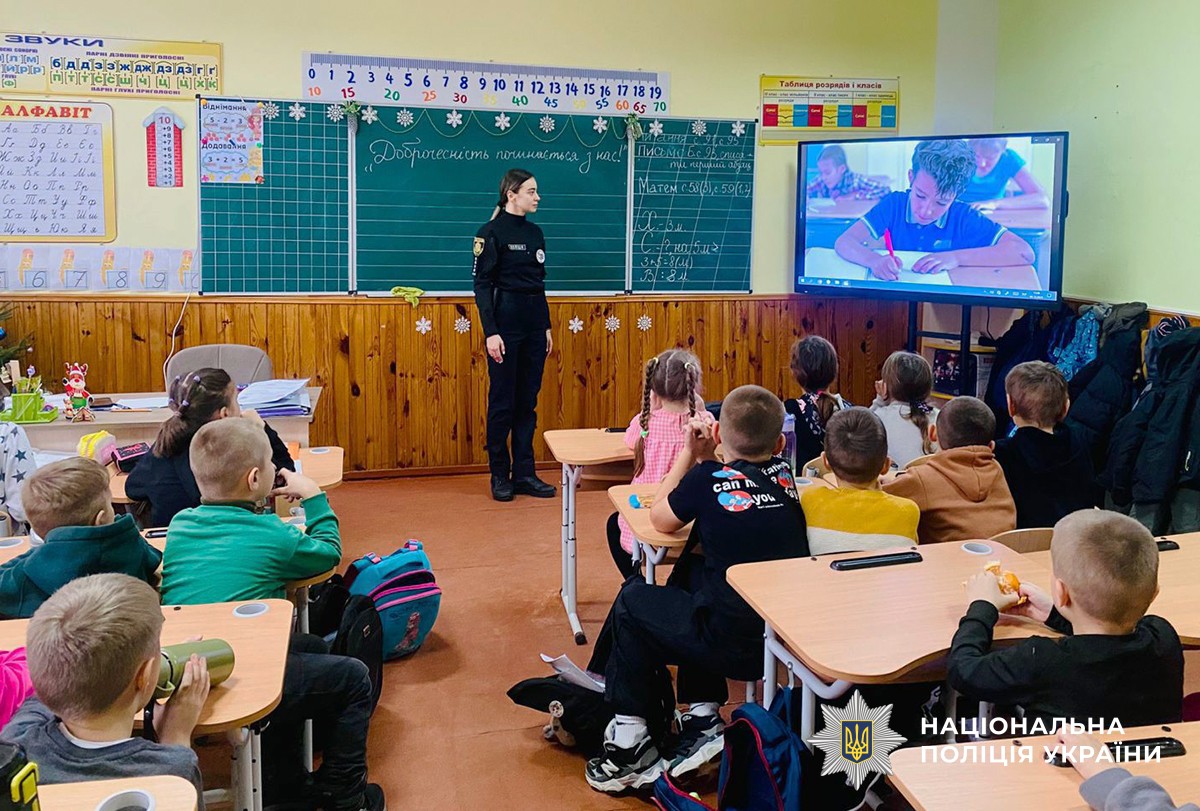 У шкількому класі, діти сидять за партами, біля зеленої шкільної дошки стоїться поліцейська, усі дивляться на екран з демонтрацією відеоролика.