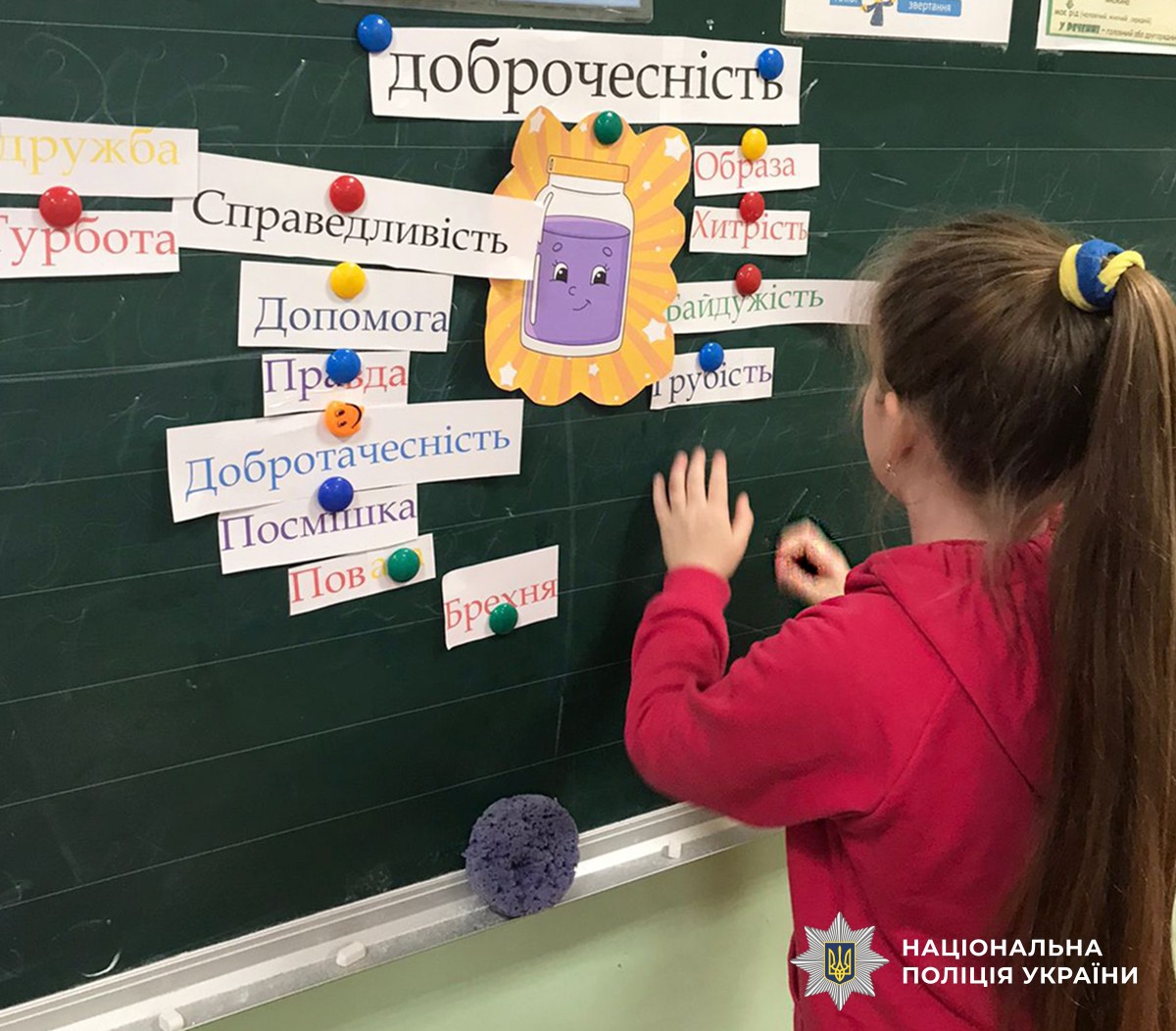 Дівчинка у рожевій кофтинці прикріплює на селену скільну дошку карточку зі словом, яке стосується доброчесності. Також вісять й інші карточки з іншими словами, що свідчить про те, що інші діти вже прикріпили.