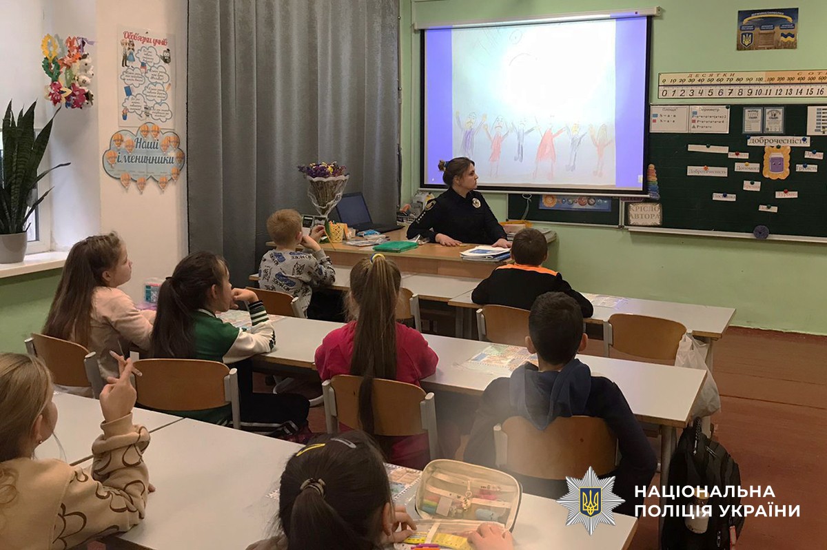 У шкількому класі, діти сидять за партами, навпроти сидить поліцейська за столом вчителя, усі дивляться на екран з демонтрацією відеоролика.